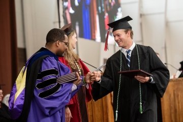 A Saint Lawrence graduate, wearing a black cap and gown, is congratulated by President Kathryn Morris and Hagi Bradley on the Commencement stage.