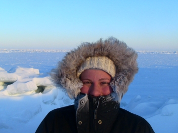 A researcher, wearing a winter jacket with a snow crusted fur hood, stands on the edge of a body of water, surrounded by snow.
