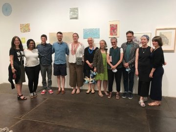 A group of people stand together at an art exhibition. Several colorful prints are on display on the wall in the background.