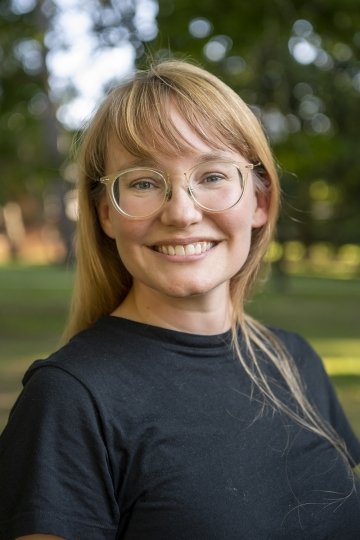 Headshot of Stacey Olney-LaPierre. She has clear-framed glasses, bangs, and is standing against a backdrop of trees and wearing a simple black dress.