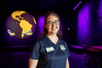 Elise Pierson '24, wearing a name badge and a blue Wild Center t shirt, stands in front of an exhibit featuring a glowing globe.