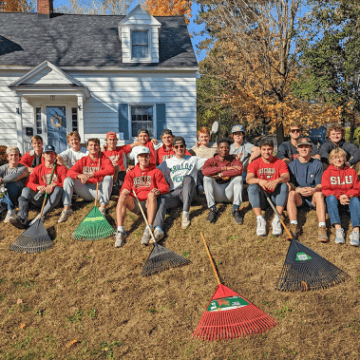 Saint Lawrence men's lacrosse team, wearing scarlet hoodies, sit on the grass outside of a home after raking leave on a sunny day.