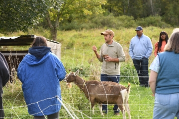 The director of the Sustainability Program gives a tour of the local farm. A brown goat stands nearby as people listen in.
