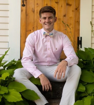 Photo of Keenan Wilson sitting on the front steps on his house. Wearing a pink button-down, long-sleeve shirt and bow tie and khaki pants.