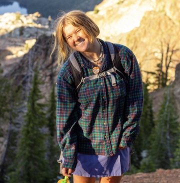 Photo of Madison Mercier smiling on the summit of a mountain wearing a green and red plaid shirt and a purple skort.