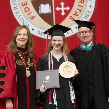 Kathryn Morris, Jacqueline Boutilier, and Michael Ranger, wearing commencement regalia, stand together on stage in celebration of Jacqueline's academic achievement award. A scarlet banner with the Saint Lawrence logo hangs in the background.