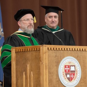 Andrew Williams, wearing green academic regalia, stands at a podium and addresses the graduating class. 