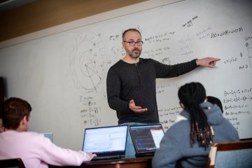 A statistics professor, wearing glasses and a dark grey henley t-shirt, stands in front of his class and points to calculations on the white board.