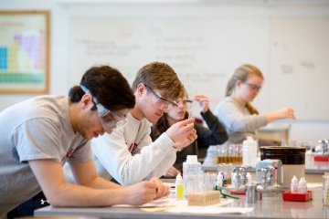 Four students sit in a row at a lab bench in chemistry class. They all wear goggles and closely study the bottles and samples in front of them. 