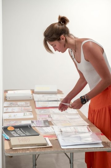 Ellie Shaw stands over a table sorting stickers and research material in a well-lit gallery with blank white walls.