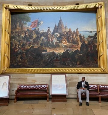 Fousani Hamidou, wearing business attire, sits on a leather bench in front of a large painting with a guilded frame in the rotunda of the U.S. Capitol Building.
