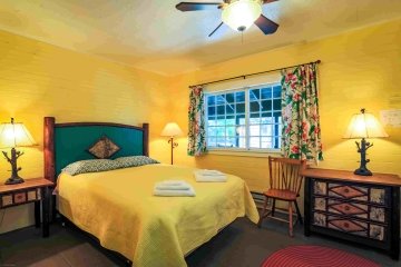 A canary yellow bedroom with a queen size bed with a turquoise headboard. Adirondack style lamps are list on Adirondack style end tables. Floral curtains hang in front of a large window. 