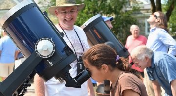 A child peers into a large black telescope as an astronomer smiles at her disovery. People in the background also peer through telescopes while one observer, wearing eclipse glasses, peers up to the sky.