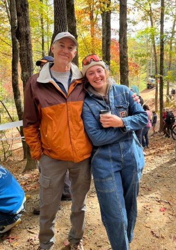 Amanda Vansant and her dad at the National Collegiate Mountain Biking Championships. Amanda wears a jean jumpsuit. Her dad has his arm around her and wears and orange windbreaker.