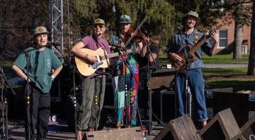 Alt text: Four musicians perform on an outdoor stage with microphones and instruments, including a guitar, bass, and violin, while smiling and wearing matching hats.