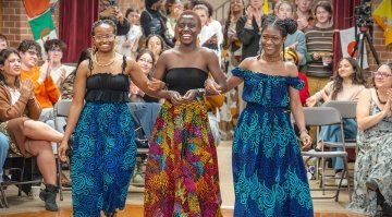 Three people wearing colorful patterned outfits walk arm-in-arm down a runway as the audience claps and smiles around them.