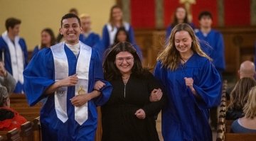 Three smiling students walk arm in arm down an aisle, with two wearing blue graduation gowns and the middle person in black.