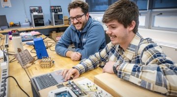 Two people in a makerspace work on a laptop, surrounded by electronics and small wooden prototypes. One person is pointing at the screen while both smile, engaged in the project. The space includes 3D printers, tools, and wiring on a long wooden table.