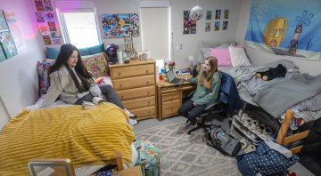 Two students sit in a cozy Saint Lawrence University dorm room. One reads on a bed with a yellow blanket, while the other works on a laptop at a desk. The room is decorated with posters, colorful bedding, and personal items.