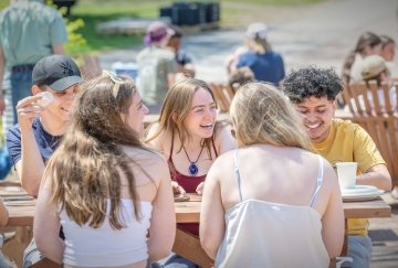 A group of students sits at a picnic table outside, smiling and laughing together on a sunny day.