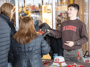 A student wearing a Saint Lawrence sweatshirt hands out materials at an indoor table while speaking with visitors.