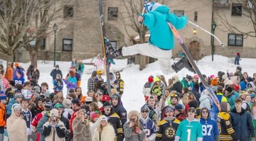 A skier performs a jump in mid-air above a large crowd of people dressed in colorful winter clothing during a snowy campus event.