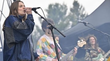 A band performs on an outdoor stage, with a singer at the microphone and musicians playing guitar and bass in the background.