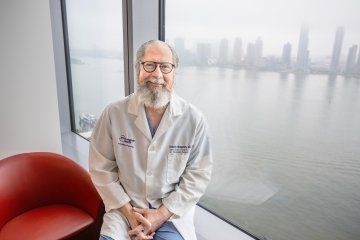 Doctor Robert Montgomery, wearing a white lab coat and blue scrubs, sits next to a large window that overlooks New York City on a rainy day.