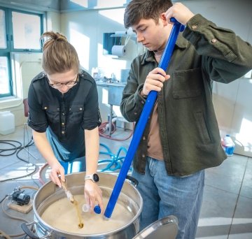 Two Saint Lawrence students stand over a large vat filled with beer they are brewing. One student tests the liquid while the other stirs with a long blue stick.