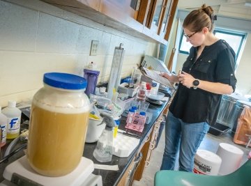 A person wearing glasses and a black shirt reviews notes on a clipboard in a science lab, surrounded by lab equipment and sample containers.