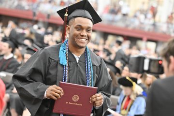 Smiling graduate in a black cap and gown holds a Saint Lawrence University diploma during a commencement ceremony.