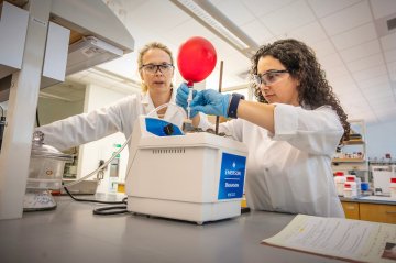 Two people in a science lab at Saint Lawrence University wear lab coats, gloves, and safety glasses while operating a benchtop instrument. One person holds tubing connected to a red balloon as they work together to monitor the experiment.
