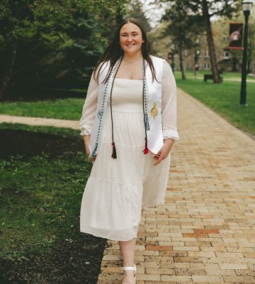 Sara St. Clair walking on a brick path wearing a white dress and graduation cords.