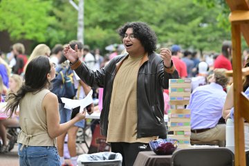 Two people chat and smile at an outdoor campus event, with one person gesturing enthusiastically beside a giant Jenga game while a crowd gathers in the background. The scene highlights a lively, social atmosphere with activities and conversation