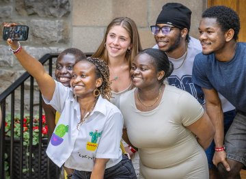 Six students smile as they gather for a group selfie outside a campus building, with one student holding up a phone to take the photo.