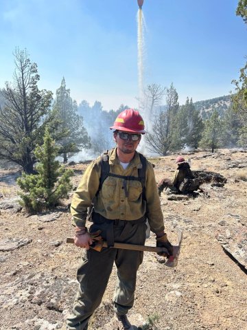 A firefighter holds a hand tool as water drops from an aircraft onto a smoky, wooded landscape behind them.