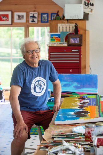 Woody Jackson sits in his art studio with vibrant paintings around him.