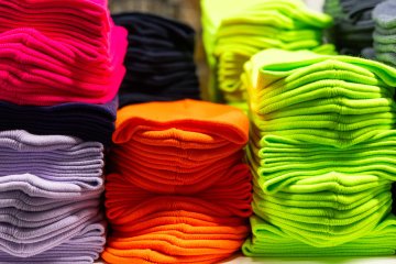 Neatly folded stacks of colorful knit clothing in bright pink, orange, neon green, purple, and black arranged in rows.