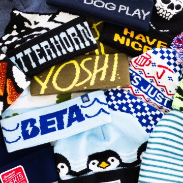 Colorful knit winter hats with bold lettering, playful patterns, and designs featuring penguins, skulls, and geometric shapes overlap in a vibrant display.