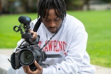 Kaleb Davis, wearing a Saint Lawrence sweatshirt, looks at the digital display of a large video camera. 