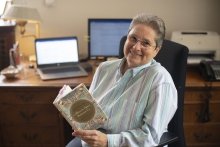 A photo of Reverend Kathleen Buckley sitting and smiling in front of her desk with a book in her hands.