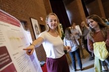 A photo of a student pointing at their research poster and presenting to an attentive listener.