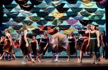 A photo of St Lawrence dancers performing on stage. They are all wearing black dresses and are dancing in front of a wall full of umbrellas.