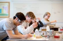 A saint lawrence student, wearing protective goggles, holds up a glass beaker and conducts research in a laboratory. 