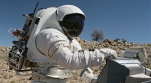 Dean Epler, wearing a NASA space suit, works on equipment in the desert. 