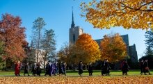 Several people wearing graduation regalia, walk in a line on a bright fall day. There are piles of orange and yellow leaves on the ground. Gunnison Memorial Chapel is in the background.