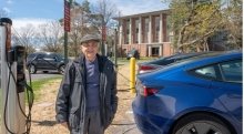 Harvey Meer, wearing a fall jacket and hat, proudly stands in front of an electric vehicle charging station on a crisp fall day. Two electric vehicles are charging behind him.