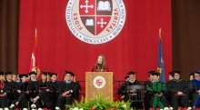 President Kathryn Morris, wearing Commencement regalia, stands behind a podium and addresses the crowd at Commencement. A large scarlet and brown banner with the Saint Lawrence crest hangs in the background.