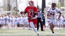 Chris Jordan, wearing a scarlet lacrosse jersey, cradles the ball away from the competition on the lacrosse field. A large group of athletes, wearing white jerseys, stand out of focus in the background.