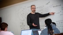 Ivan Ramler, wearing a dark grey shirt, stands in front of a white board and points at a math problem. Students, sitting in front of laptops, listen intently to the lecture.